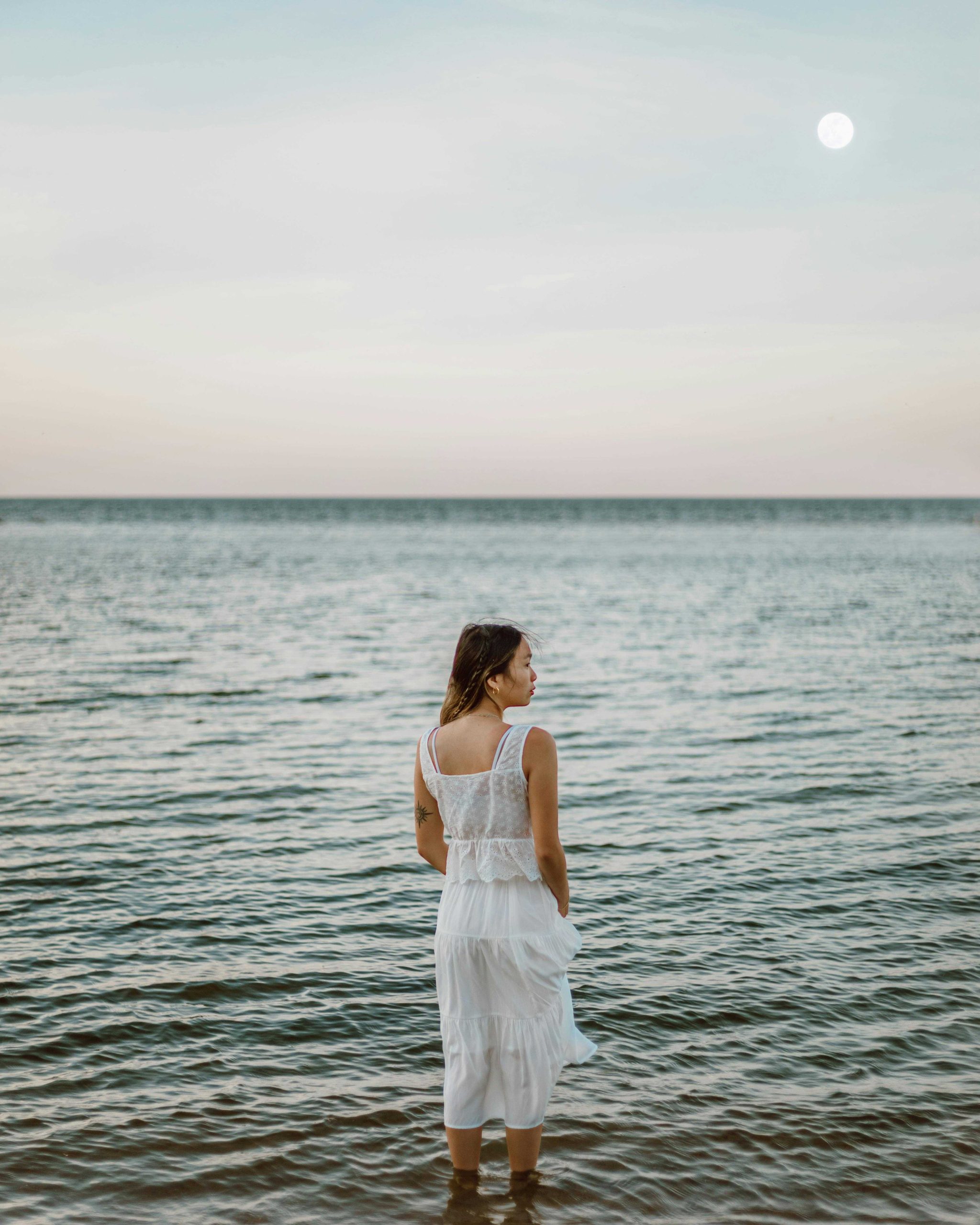 A woman stands on the seafront, gazing out over the ocean with the moon high in the sky.